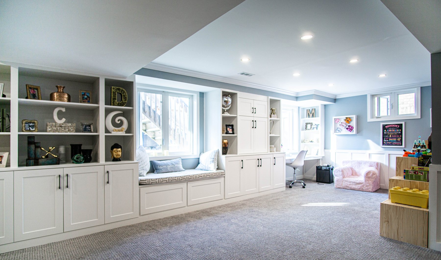 Bright finished basement with white built-in cabinets, shelving and a window seat